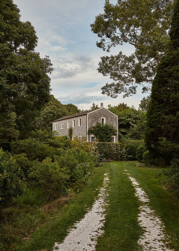 Exterior view of a grey two-story house surrounded by lush green trees and foliage, gravel driveway leading to the residence.