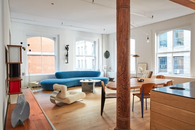 Wide interior of a bright, airy loft apartment with a curved blue sofa, modern furniture, wood floors, a wooden pillar, and large windows.