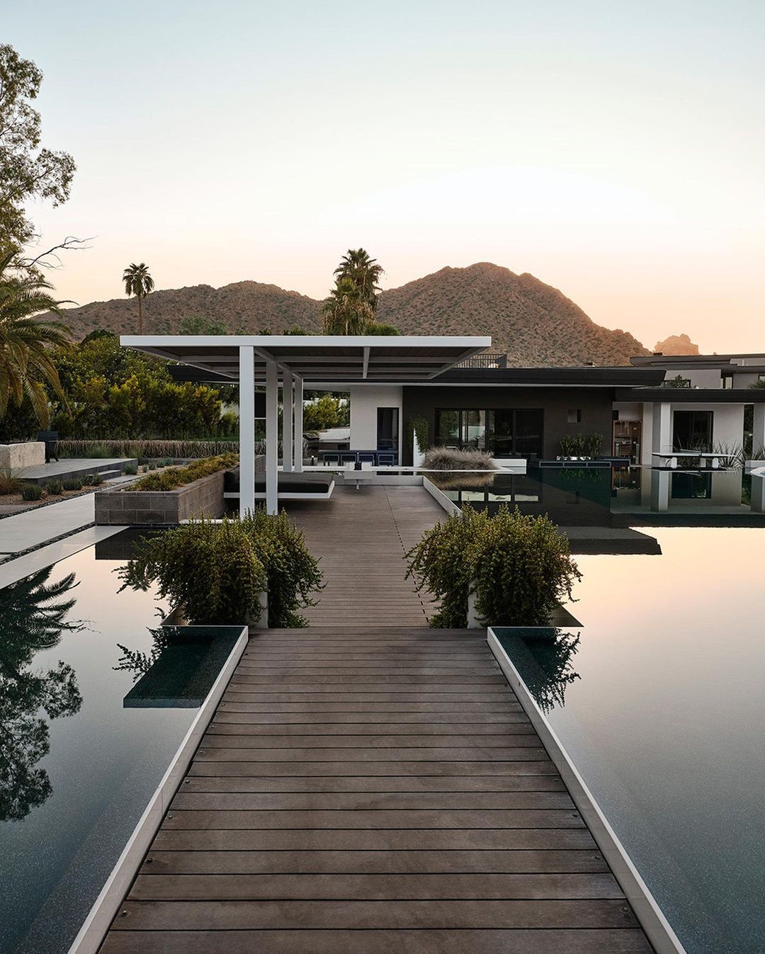 Exterior view of a modern house with a pool, deck, and mountain backdrop at sunset, showcasing architectural design and landscaping.