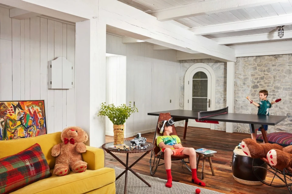 Indoor playroom scene: a boy wearing a VR headset sits, another plays table tennis, and a yellow couch features a teddy bear.