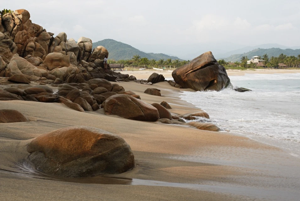 Tranquil beach scene with rock formations along the shoreline, meeting gentle ocean waves under a soft sky, with distant mountains.