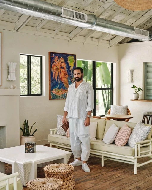 Man with a beard stands in a bright living room with a painting, white furniture, woven stools, and neutral decor in a home interior setting.