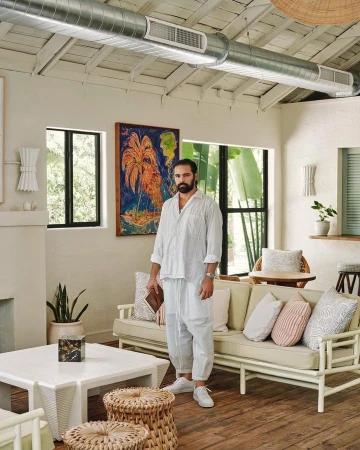 Man with a beard stands in a bright living room with a painting, white furniture, woven stools, and neutral decor in a home interior setting.