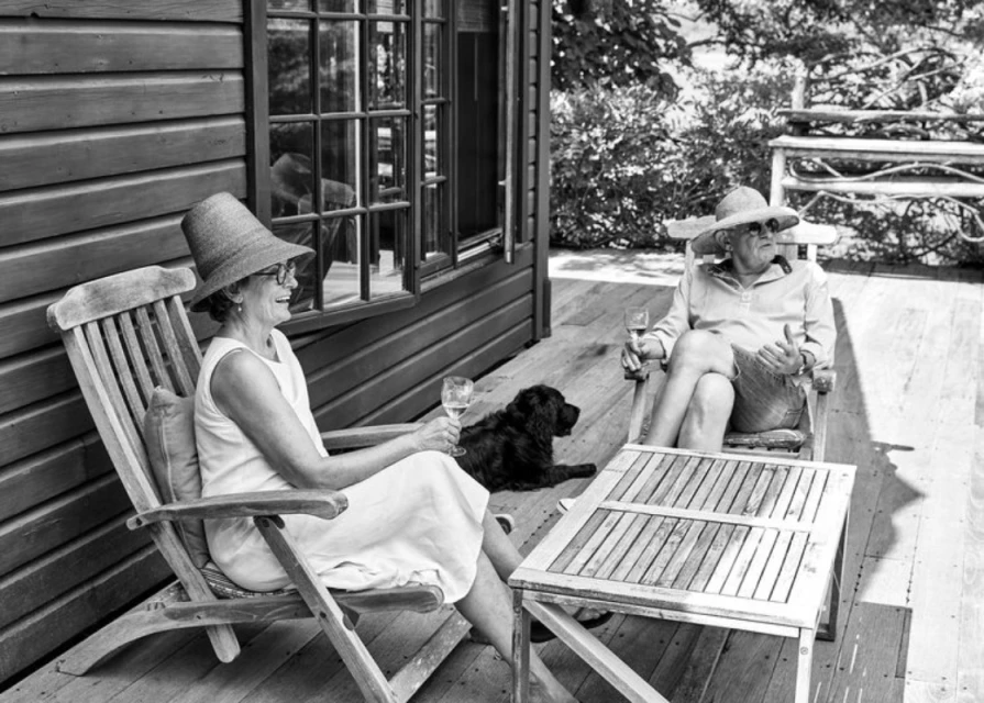 Two elderly women wearing hats sit in wooden chairs outside with a dog and glasses of wine on a deck next to a wood-sided building.