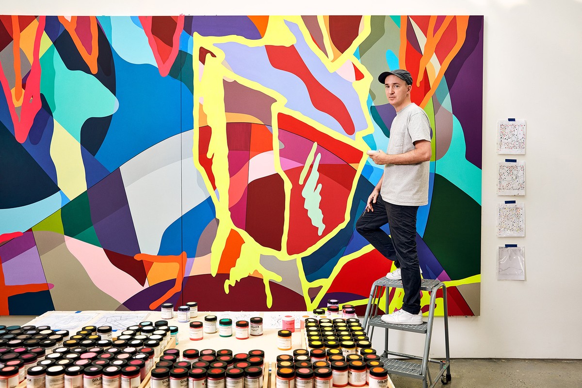 An adult male artist stands on a metal rolling ladder next to a very large, colorful, abstract painting on a white wall in his art studio.