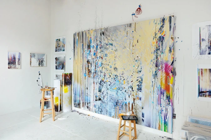 Large abstract painting displayed in a bright art studio, featuring a variety of colors and textures, with art supplies nearby.