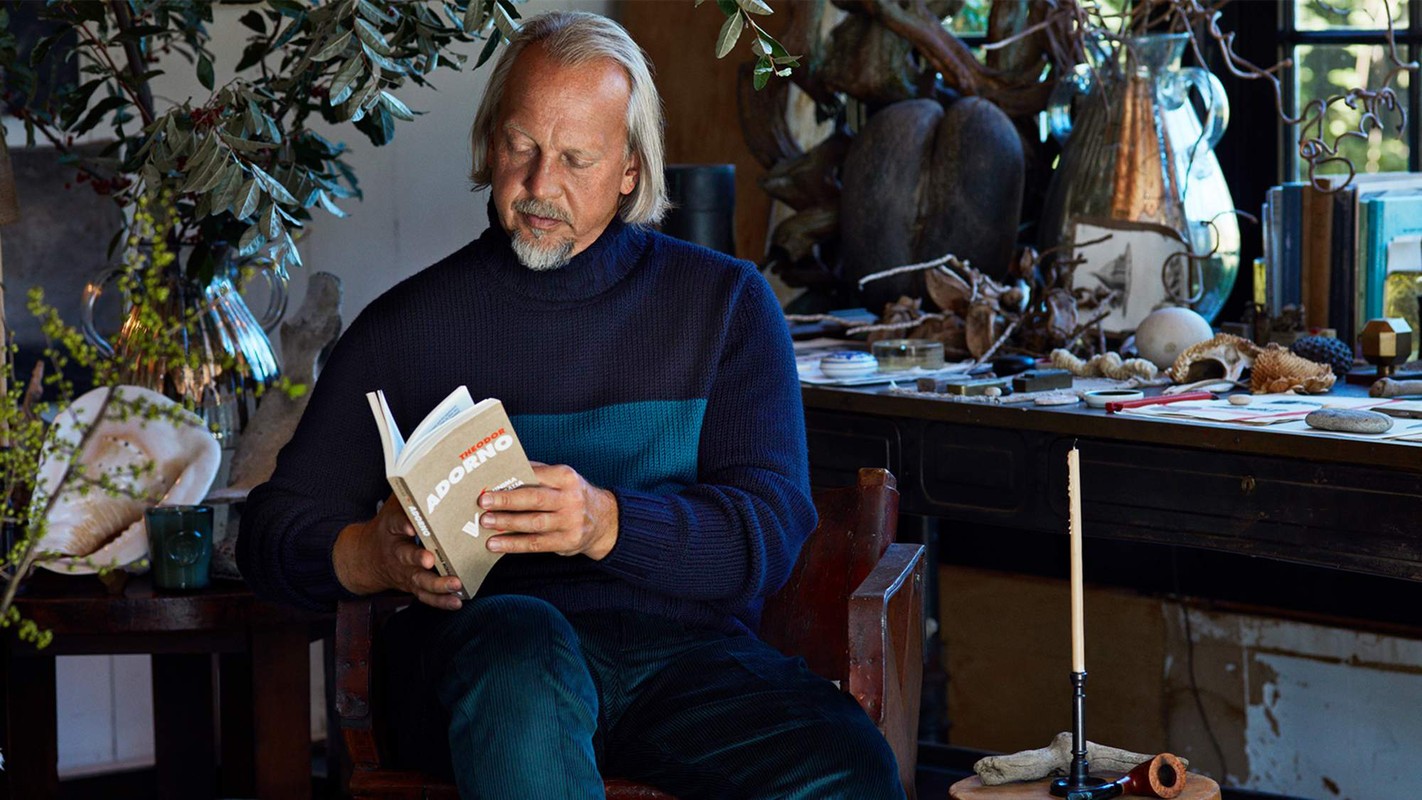 A man with grey hair and beard sits in a chair, reading a book in a room with plants and various objects on a desk in the background.