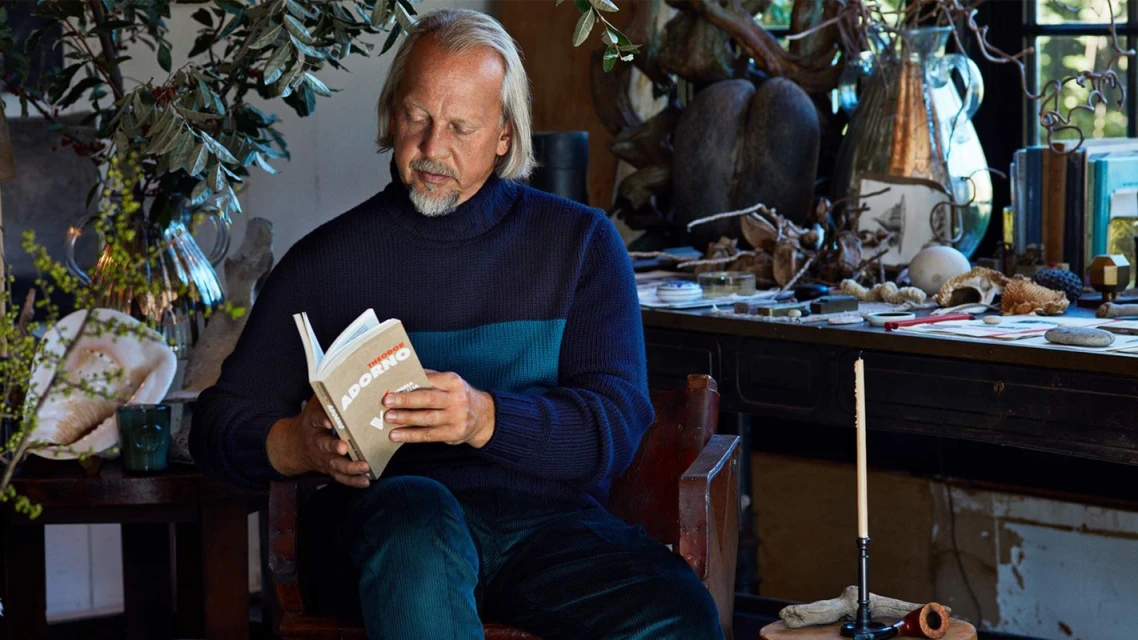 A man with grey hair and beard sits in a chair, reading a book in a room with plants and various objects on a desk in the background.