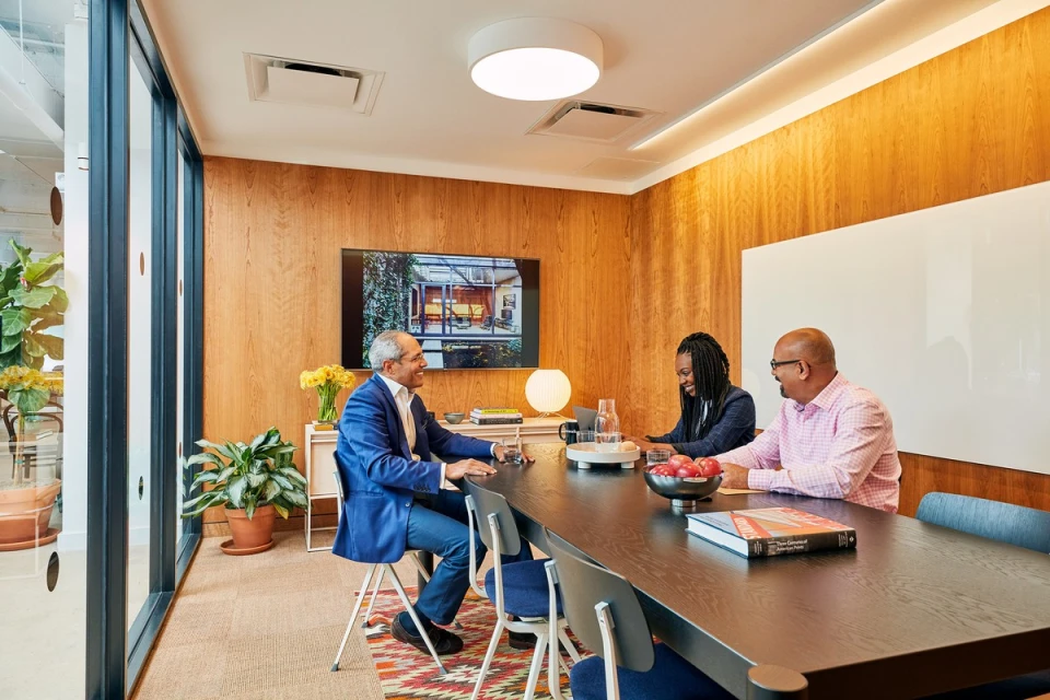 Three adults at a business meeting around a dark wood table in a conference room with wood-paneled walls and modern decor and lighting.