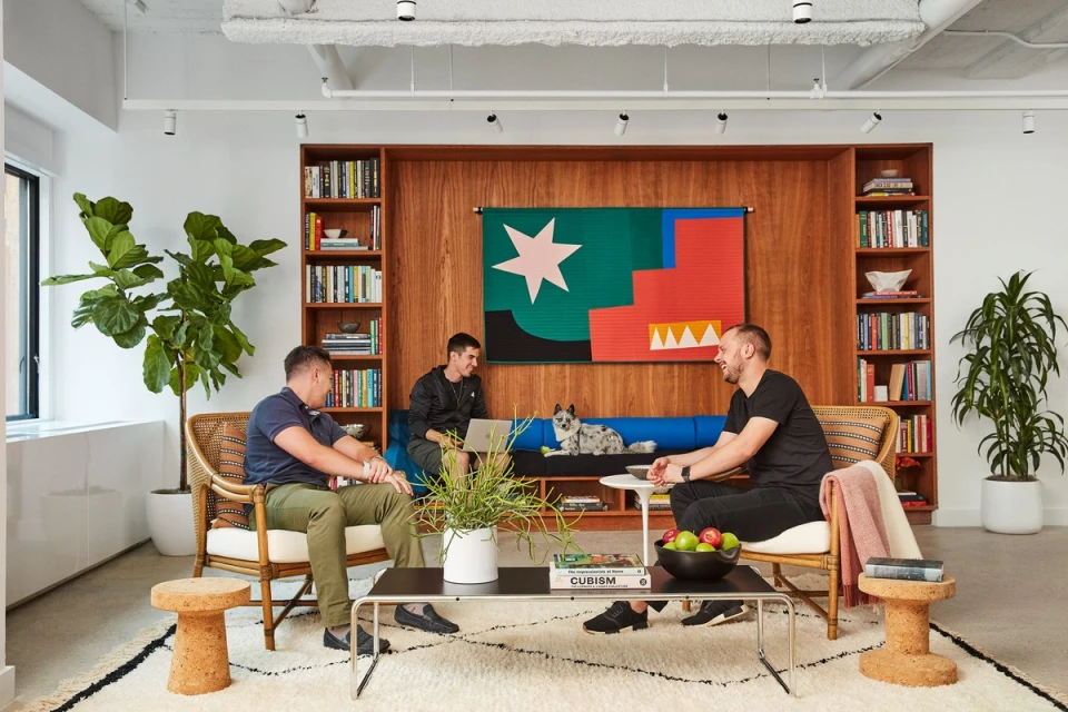 Three adult men are conversing in a modern office lounge area with a dog, surrounded by books, plants, and comfortable seating.