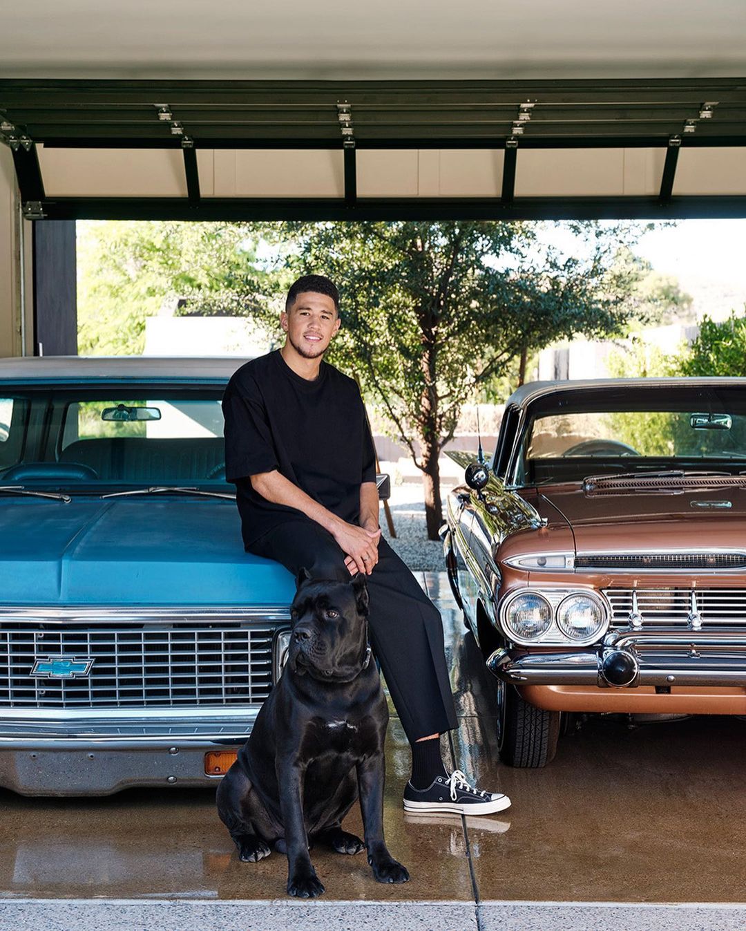 Devin Booker poses with his dog in front of classic cars, inside garage with open door to greenery. Dark teal and brown vehicles.