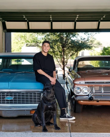 Devin Booker poses with his dog in front of classic cars, inside garage with open door to greenery. Dark teal and brown vehicles.