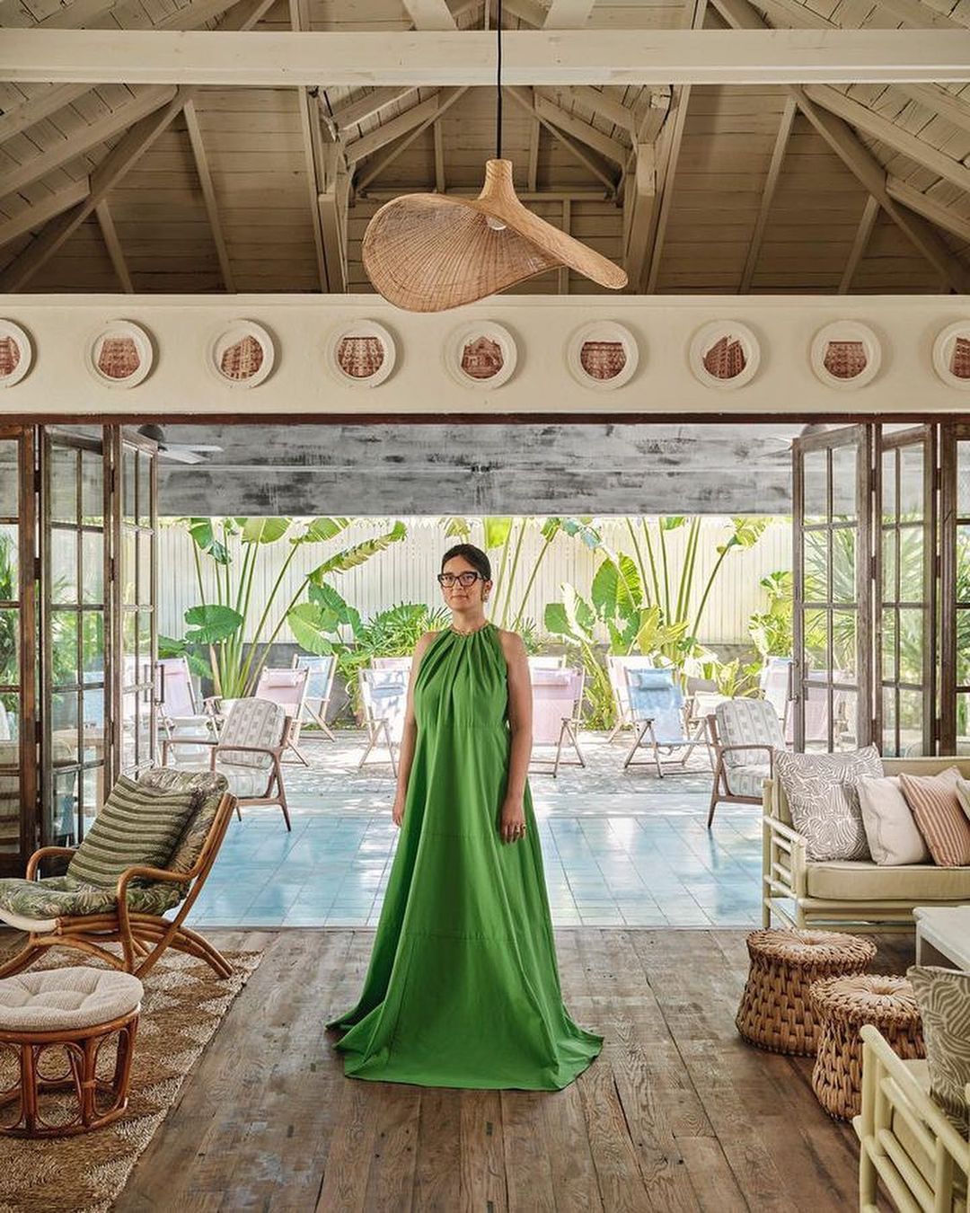 Woman in a bright green gown stands in a room with wood floors and wicker decor, looking toward the camera, with an open patio behind.