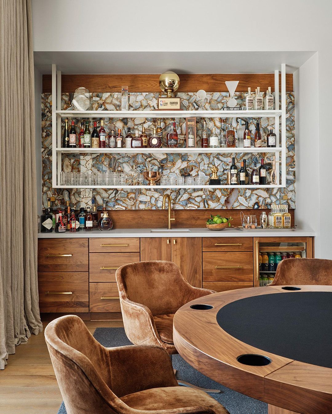 Stylish home bar setup with wooden cabinets, mirrored backsplash, open shelves, and brown velvet chairs around a poker table.