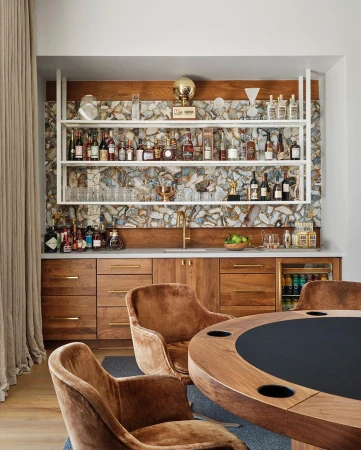 Stylish home bar setup with wooden cabinets, mirrored backsplash, open shelves, and brown velvet chairs around a poker table.