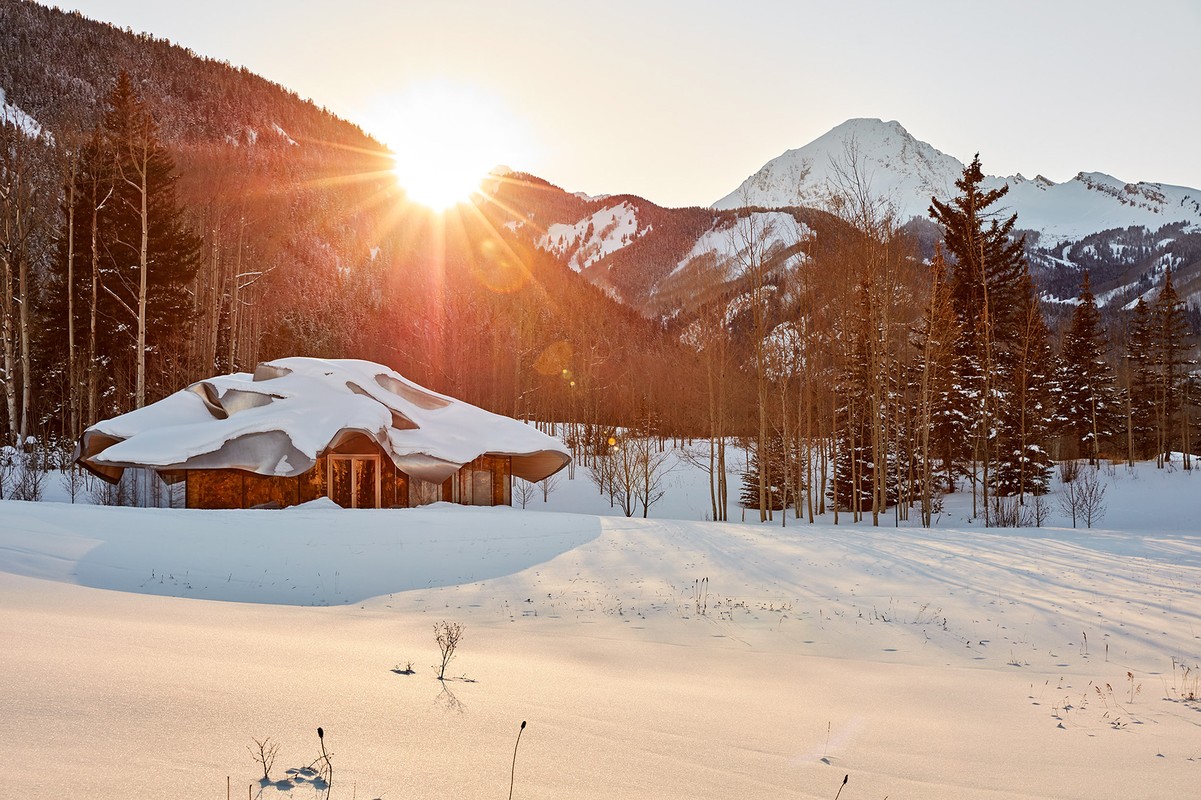 Snow-covered house in a winter landscape with mountain views. Sunlight peeks through trees illuminating the snowy ground and wooden structure.