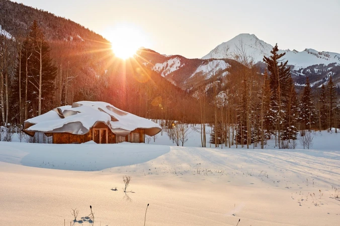 Snow-covered house in a winter landscape with mountain views. Sunlight peeks through trees illuminating the snowy ground and wooden structure.