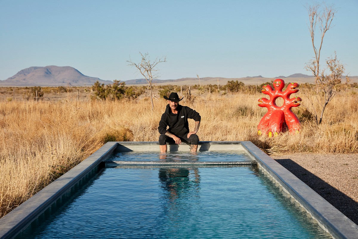 Man with cowboy hat sits at the edge of a pool in the desert with a red sculpture in the background on a clear, sunny day.