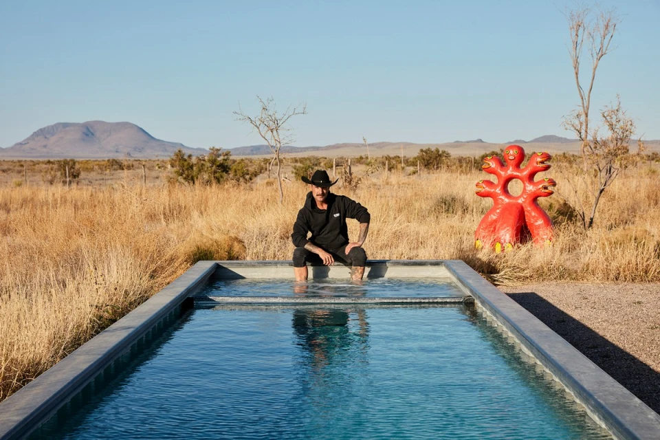 Man with cowboy hat sits at the edge of a pool in the desert with a red sculpture in the background on a clear, sunny day.