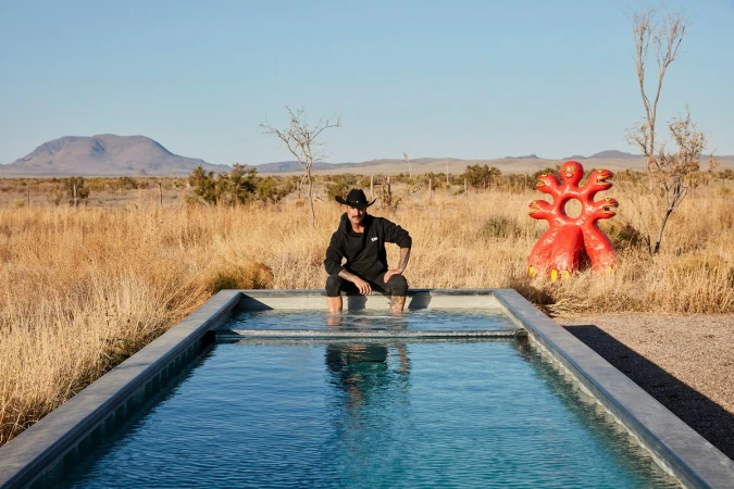 Man with cowboy hat sits at the edge of a pool in the desert with a red sculpture in the background on a clear, sunny day.