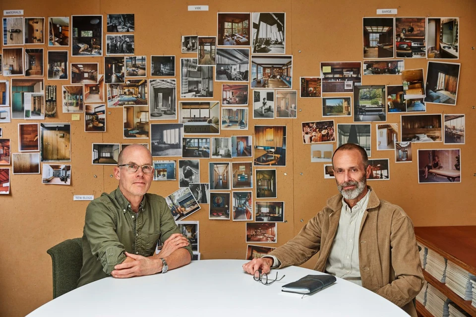 Two adult males in a meeting room with a corkboard featuring interior design photos, presenting an architectural concept in a modern style.