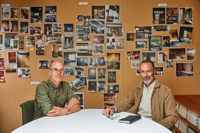 Two adult males in a meeting room with a corkboard featuring interior design photos, presenting an architectural concept in a modern style.