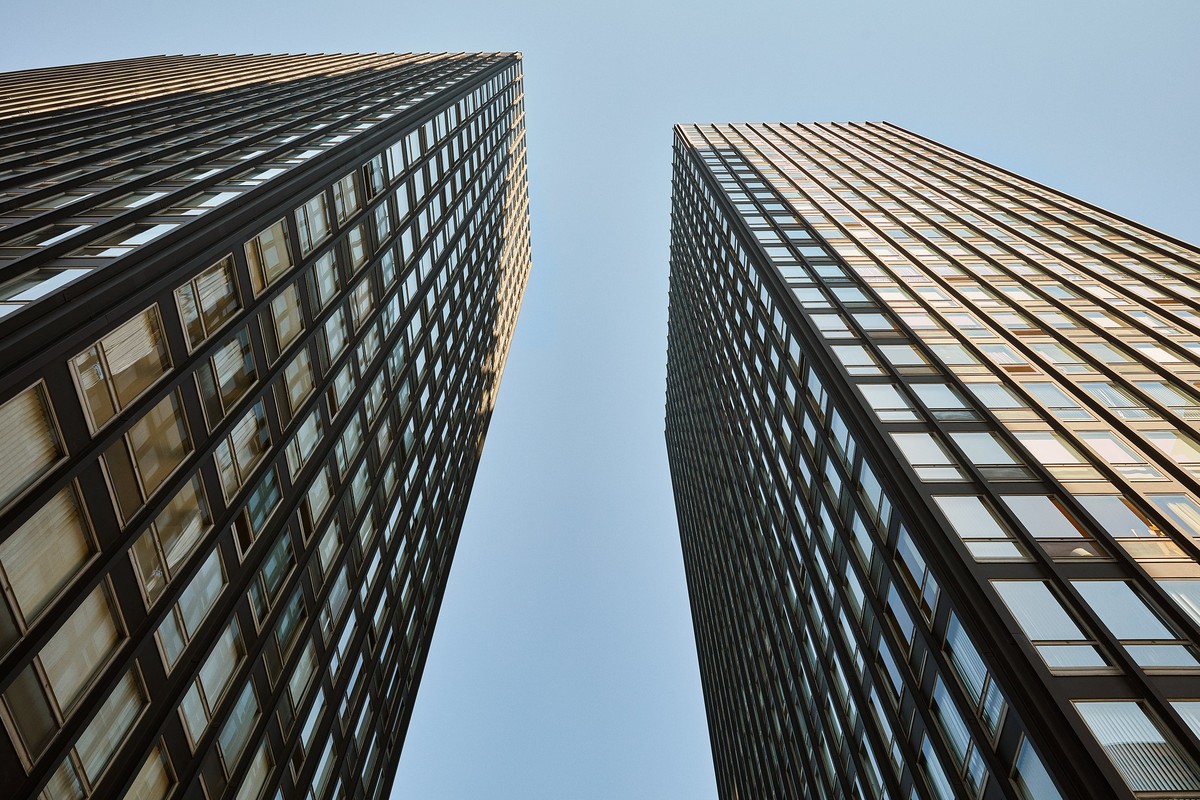 Low-angle view of two modern, dark, high-rise buildings against a light blue sky, emphasizing their height and architectural detail.