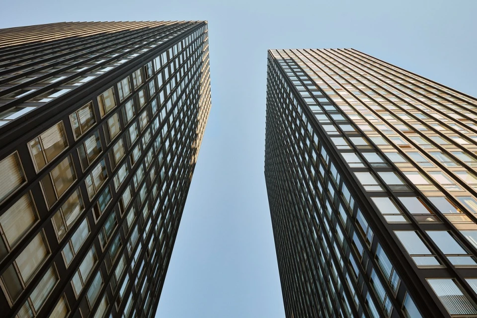 Low-angle view of two modern, dark, high-rise buildings against a light blue sky, emphasizing their height and architectural detail.