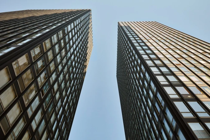 Low-angle view of two modern, dark, high-rise buildings against a light blue sky, emphasizing their height and architectural detail.