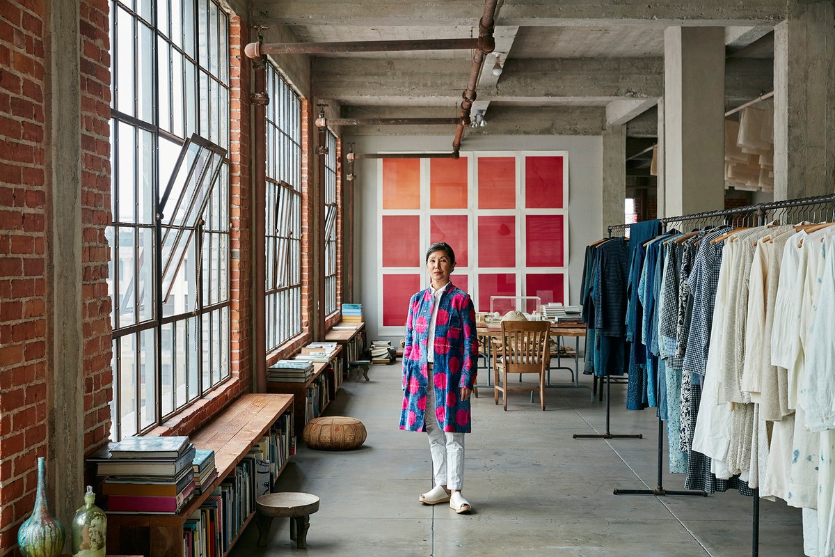 An Asian woman stands inside a converted industrial space with large windows, wearing a patterned coat, white pants, and light-colored flats.