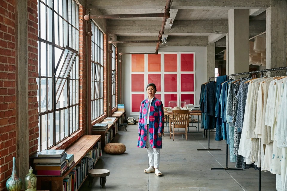 An Asian woman stands inside a converted industrial space with large windows, wearing a patterned coat, white pants, and light-colored flats.