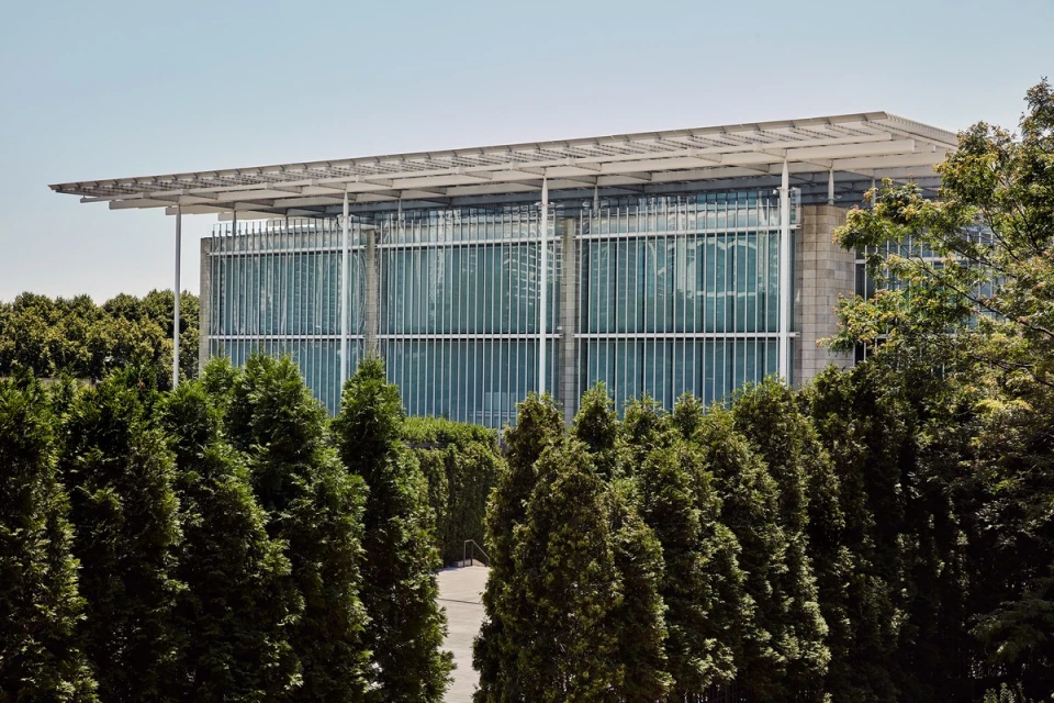 Exterior view of a modern, glass-walled building with a flat roof and steel supports, surrounded by tall, green coniferous trees under a clear sky.