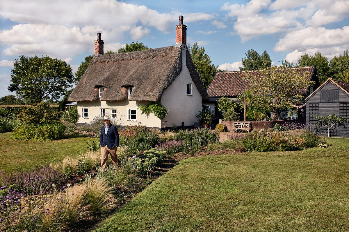 Man in blazer walks in garden path near thatched-roof cottage on sunny day; lush lawns, trees, blue sky with clouds create idyllic scene.