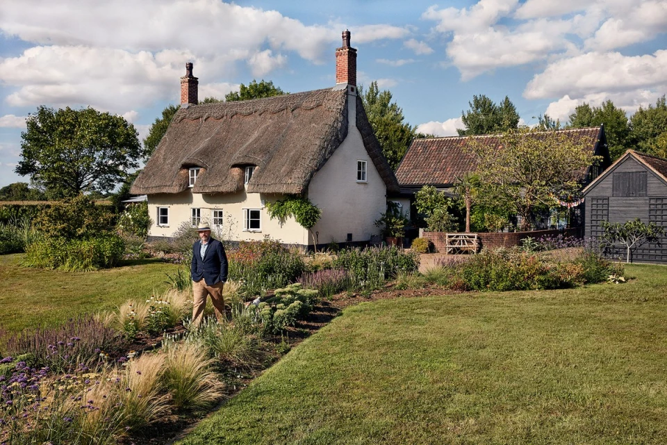 Man in blazer walks in garden path near thatched-roof cottage on sunny day; lush lawns, trees, blue sky with clouds create idyllic scene.