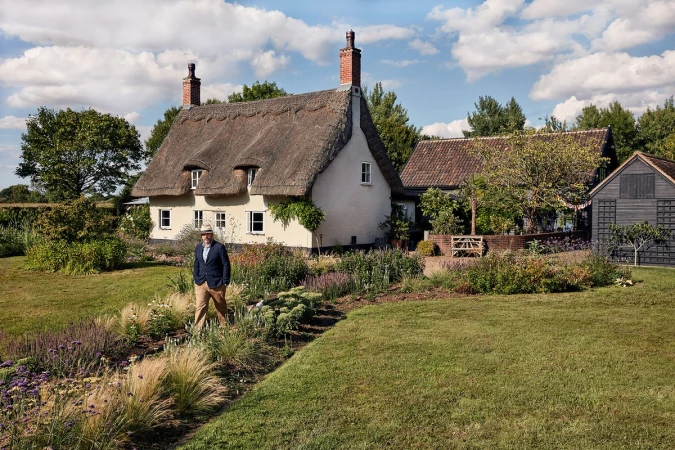Man in blazer walks in garden path near thatched-roof cottage on sunny day; lush lawns, trees, blue sky with clouds create idyllic scene.
