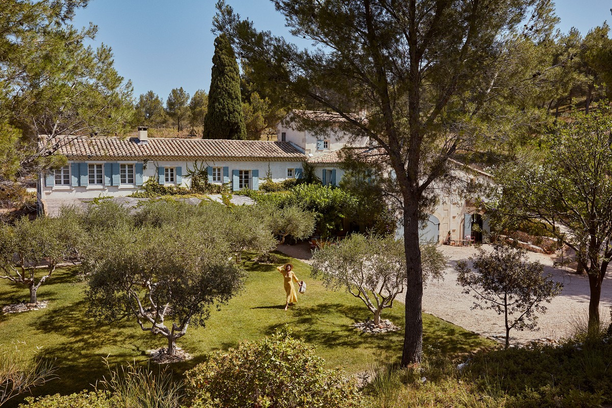 Scenic view of a woman in a yellow dress walking on the lawn of a French-style villa surrounded by trees on a sunny day.