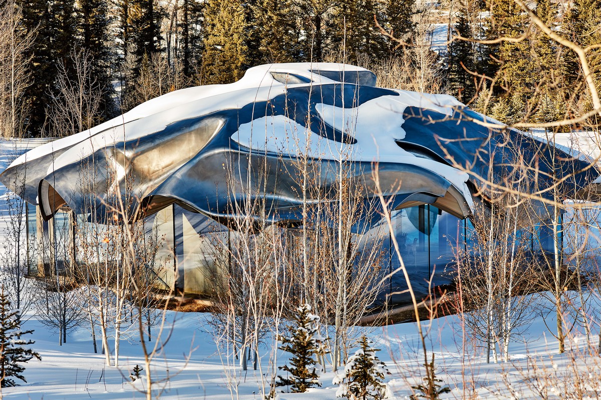Modern architectural house exterior covered in snow, with large glass windows and a reflective metal roof in a wintery forest landscape.
