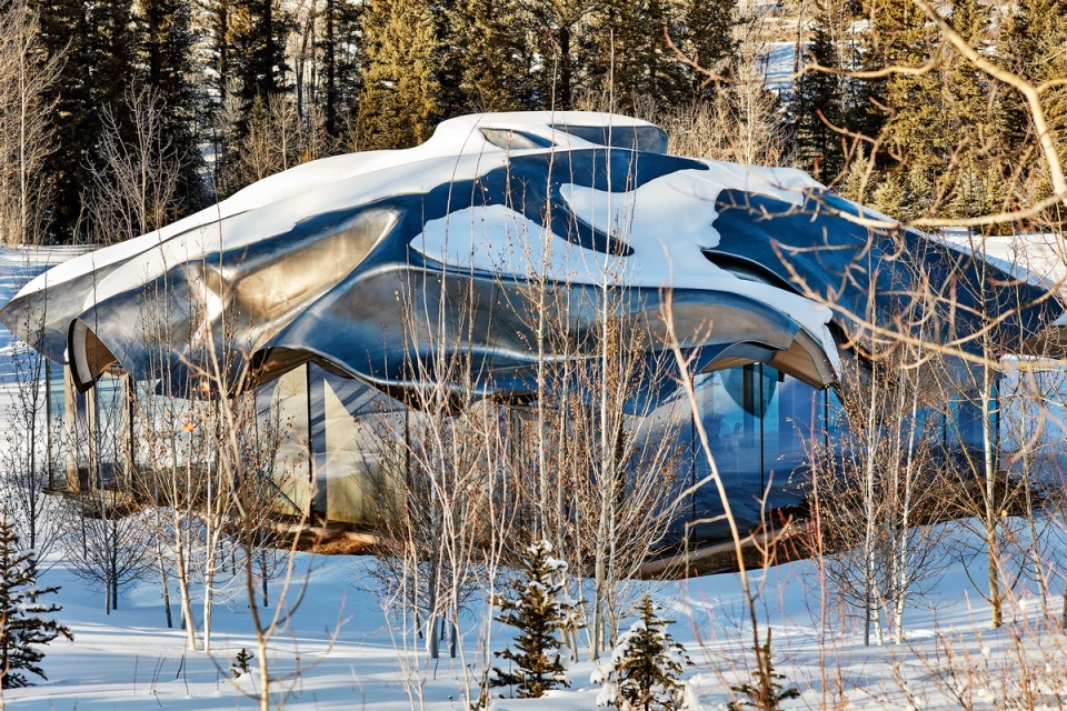 Modern architectural house exterior covered in snow, with large glass windows and a reflective metal roof in a wintery forest landscape.