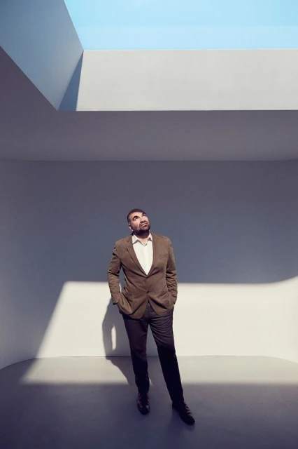 Adult man in a brown suit stands in a minimalist grey room looking up at a skylight, with sunlight and shadow accentuating the scene.