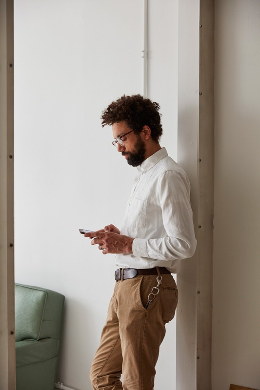Adult male with beard and glasses leaning against wall while using a phone, wearing a white shirt and khaki pants in an indoor setting.