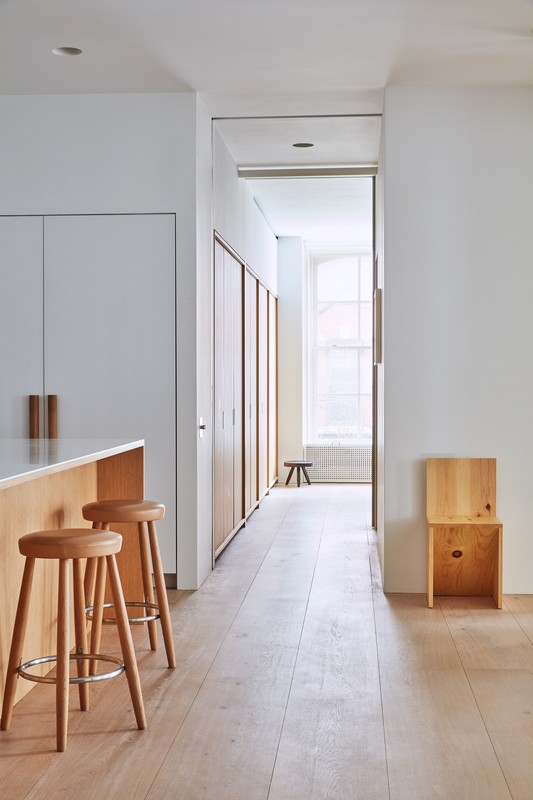 Bright interior photo of a modern kitchen and hallway featuring wooden stools, cabinets, and minimalist decor with light wood flooring.