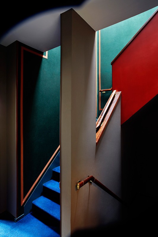 Architectural photo of a staircase featuring bright blue carpet, red wall, a wooden handrail, and green wallpaper in the background.