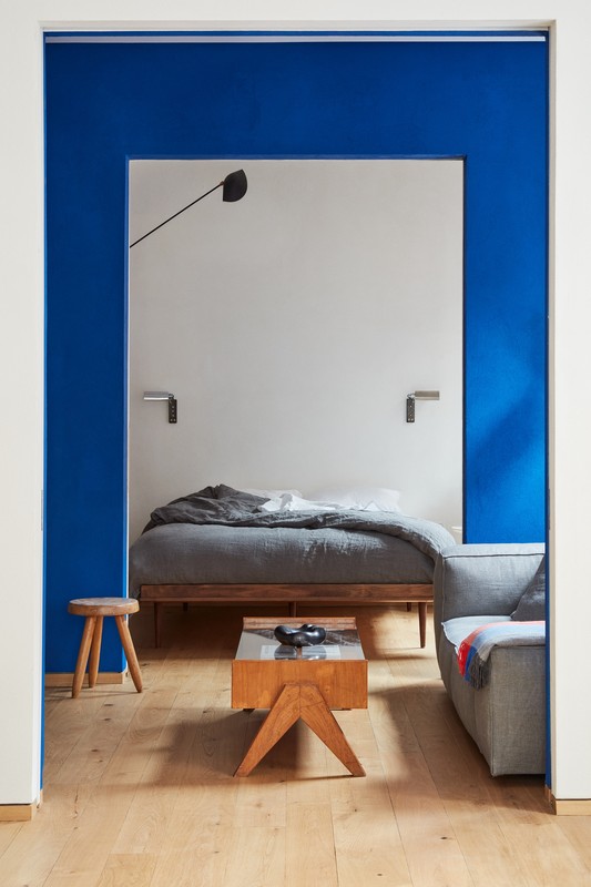 Interior view of a modern apartment showcasing a bed in a room with blue walls, visible through an opening, with a gray sofa nearby.