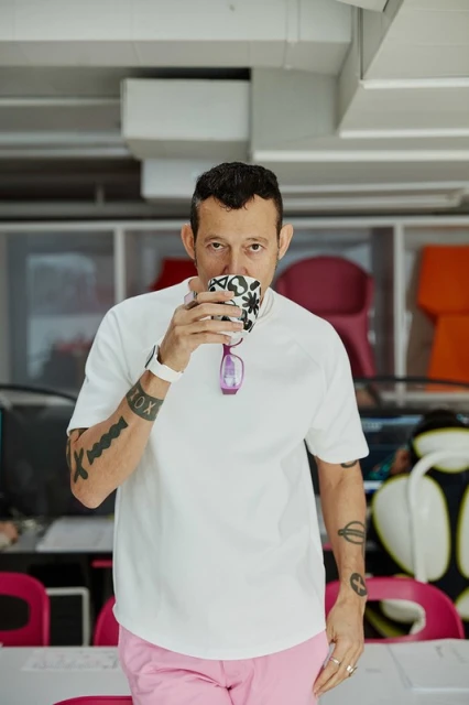 Adult man wearing a white t-shirt and pink pants drinking from a patterned mug indoors with modern office furniture in the background.