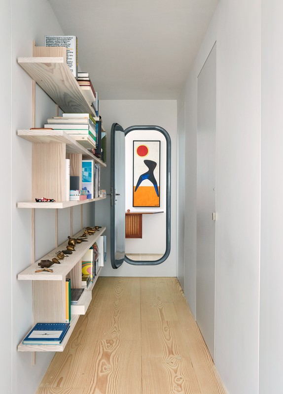 Hallway featuring a blonde wood floor, white walls, and a built-in bookshelf. A door offers a glimpse into another room with artwork.