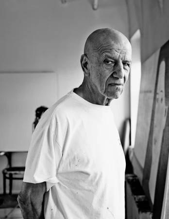 Portrait of an elderly bald man in a white t-shirt looking toward the camera in a bright studio space