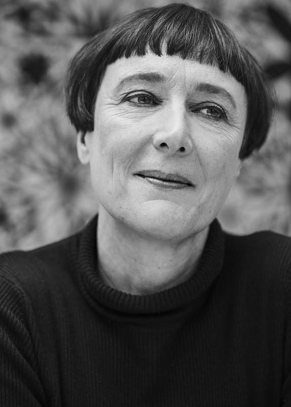 Black and white portrait of an adult female with short hair and a dark turtleneck sweater against a patterned backdrop. Head and shoulders.