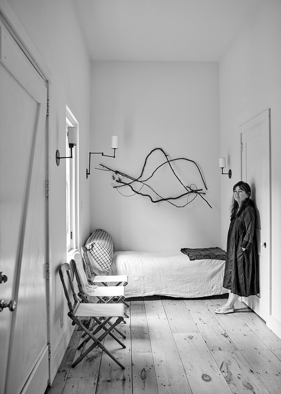 Black and white interior photo of a woman in a long dark dress, standing near a white door in a hallway with chairs and art.