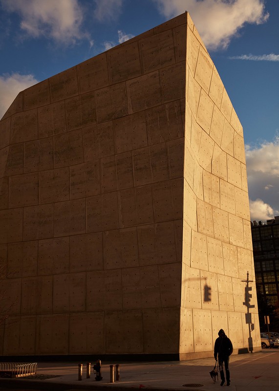 Large concrete building with an unusual shape, set against a blue sky with clouds, with a person and dog on the sidewalk.