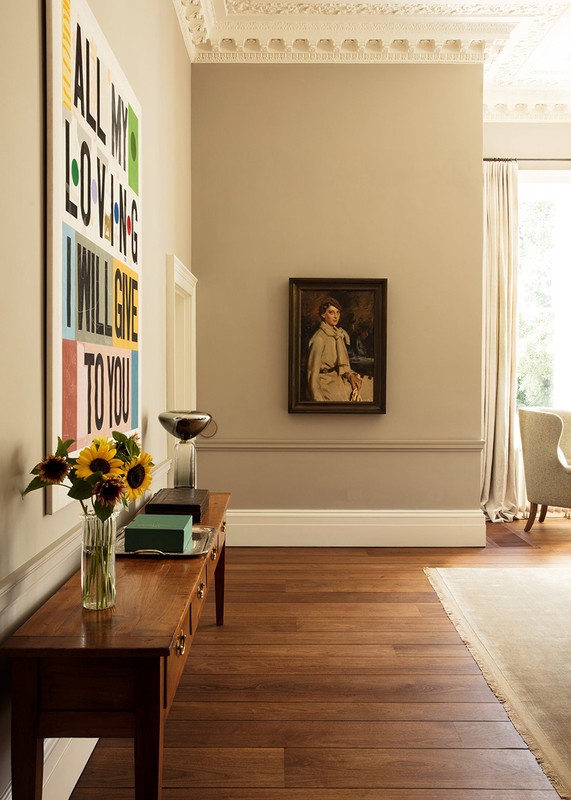 Hallway with wood floors, a console table with flowers and objects, colorful artwork, and a framed portrait on a neutral-colored wall.